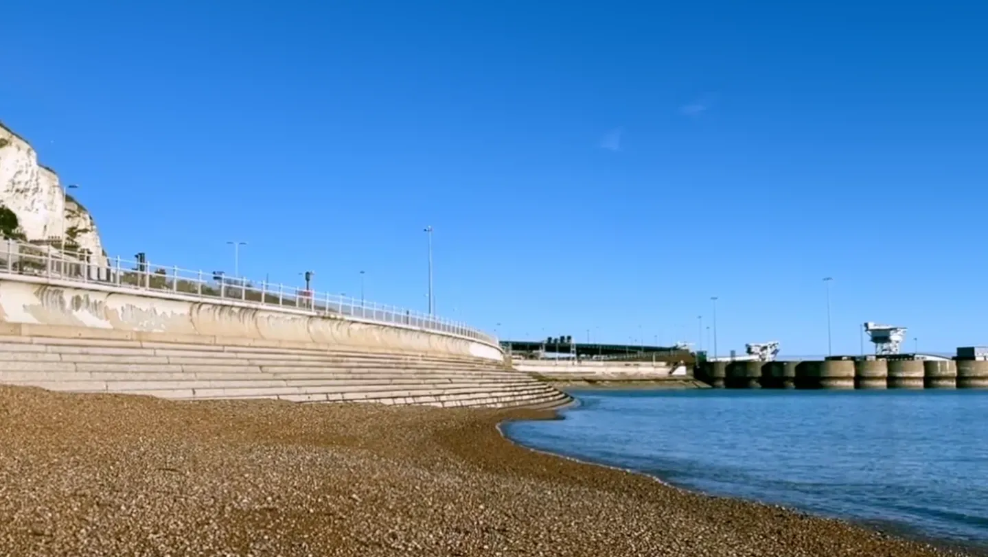 A still showing the eastern port and shingle beach at Dover, with the white cliffs to the left and the ferry port to the right