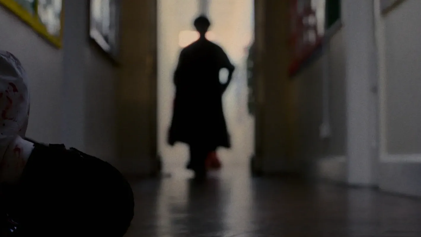 A dark figure in black approaches a wounded school student on the floor