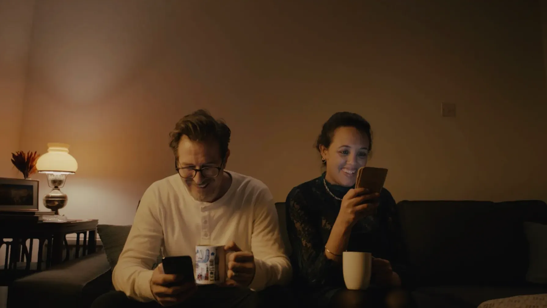 A Caucasian man in his 40s wearing glasses, and a North African woman in her 30s, both sit next to each other on a sofa with a mug in one hand and their phone in the other hand.
