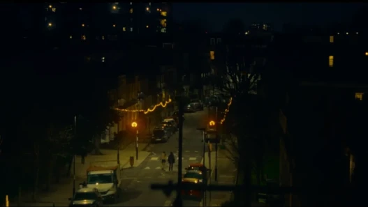 Two women walk down a dimly lit street under warm string lights at night.