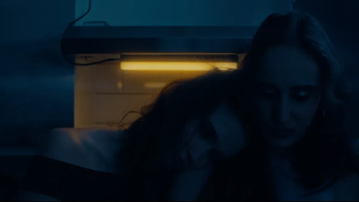 Two young women sit quietly in a dim blue-lit kitchen, one resting her head on the other's shoulder beneath a yellow light.