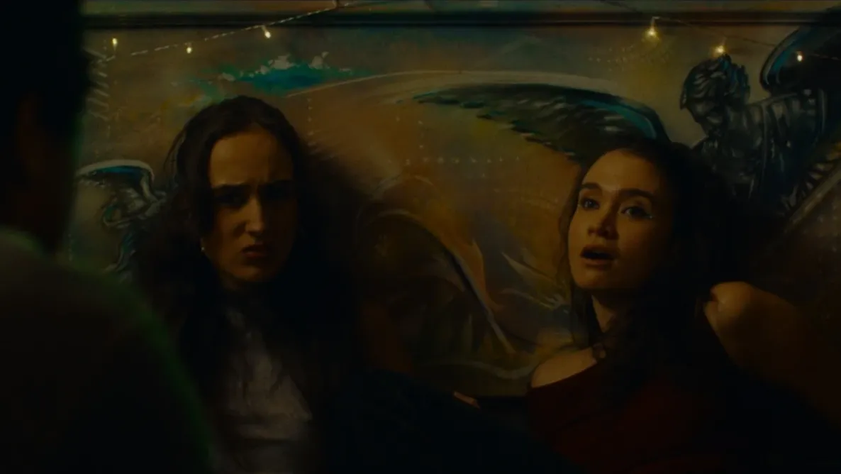 Two women sit on the floor of a pub beneath fairy lights, reacting emotionally against a mural wall.