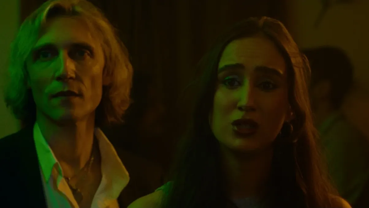 A woman looks distressed at a party, lit in green and orange hues, with a man standing tensely behind her.