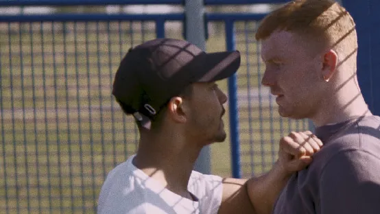 Two men in confrontation with each other, one has the other pushed against a metal sports cage