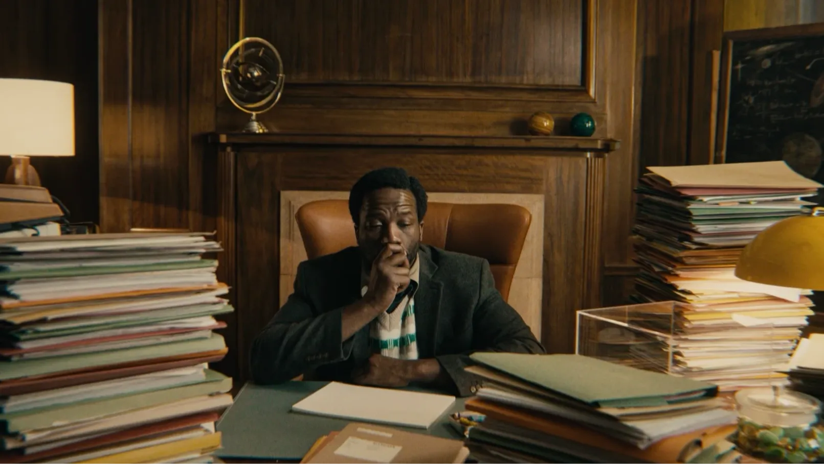 A man sitting at a desk in an office surrounded by mountainous stacks of files and paperwork.