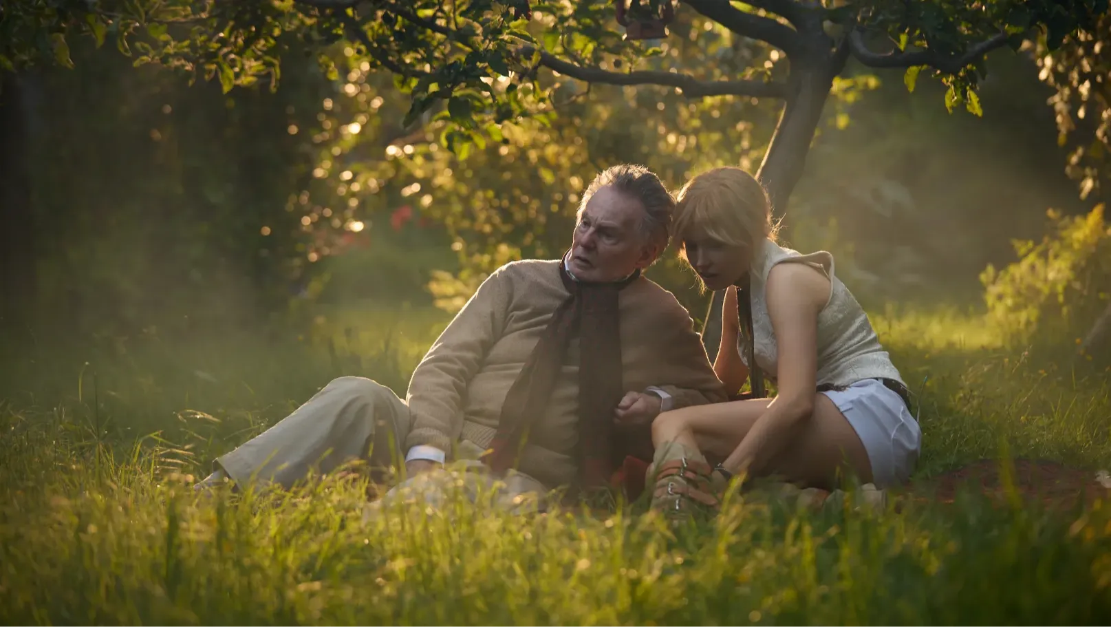An older man and a young woman sit on a blanket in a sunlit garden.