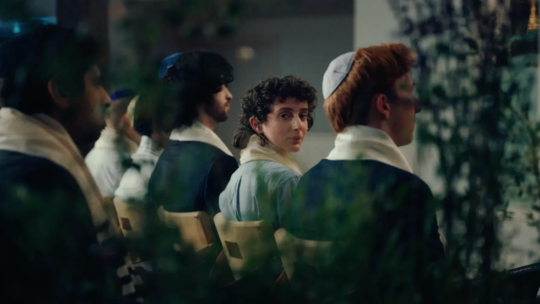 A dark haired non binary Jewish person sits amongst the men in the men's section of a synagogue.