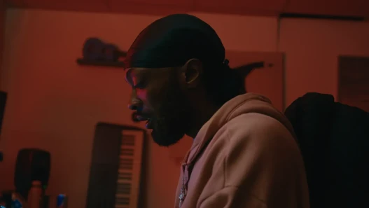 Carter the Bandit, a black man, sits in a music studio wearing a pink hoodie and black durag. There is a pink light shining on his face.