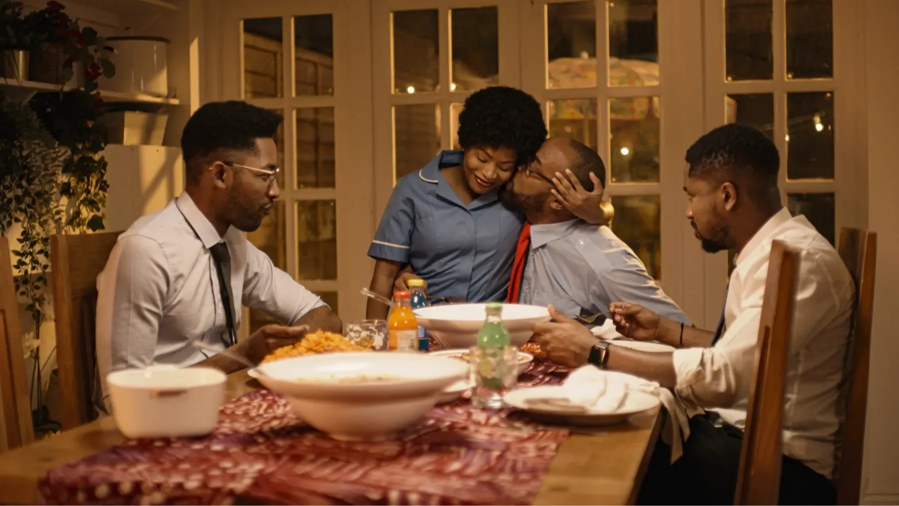 A family sitting around a dinner table with food, sharing a meal in a warmly lit dining room. One man is embracing a smiling woman dressed in blue while the children look on.