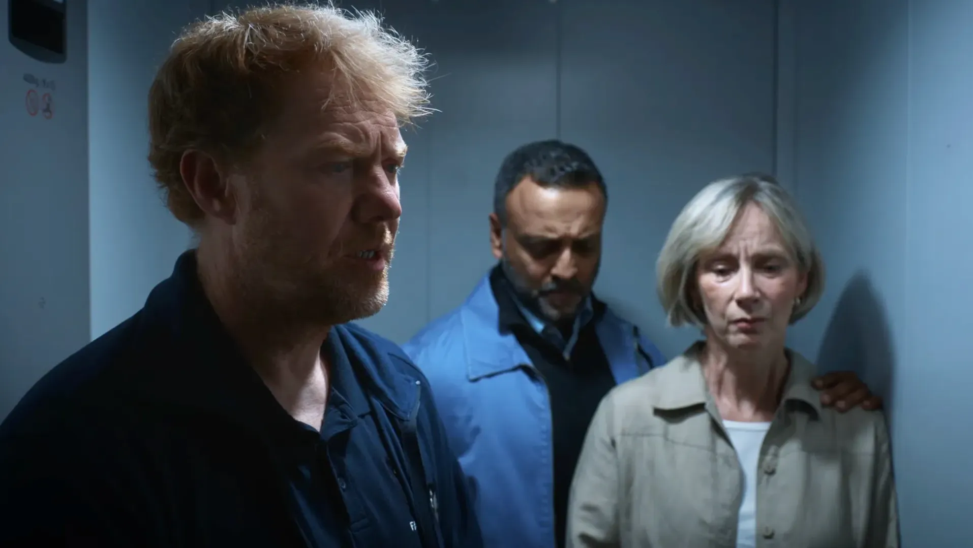 A man in a caretaker's uniform and two other adults, all looking sad, are standing in a lift.