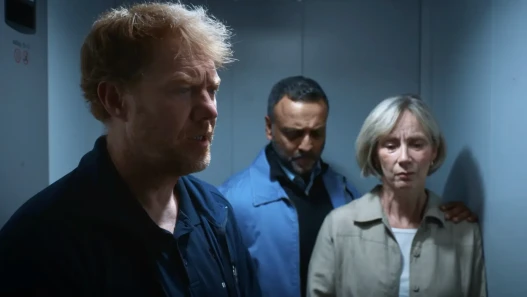 A man in a caretaker's uniform and two other adults, all looking sad, are standing in a lift.