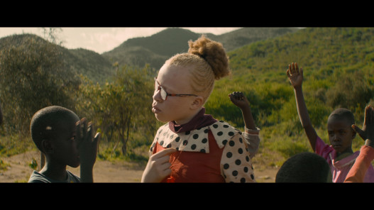 A girl wearing glasses is standing outside. She is surrounded by smaller children who have one arm raised as if answering a question.