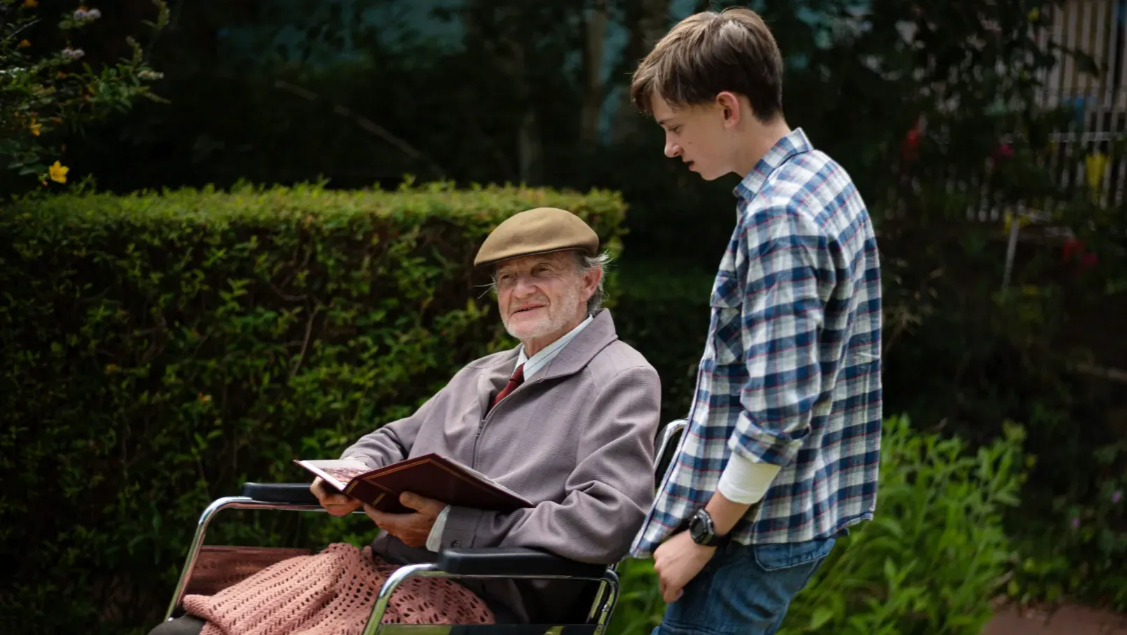 A teenage boy is standing next to a man seated in wheelchair. The man has a photo album open in his lap. 