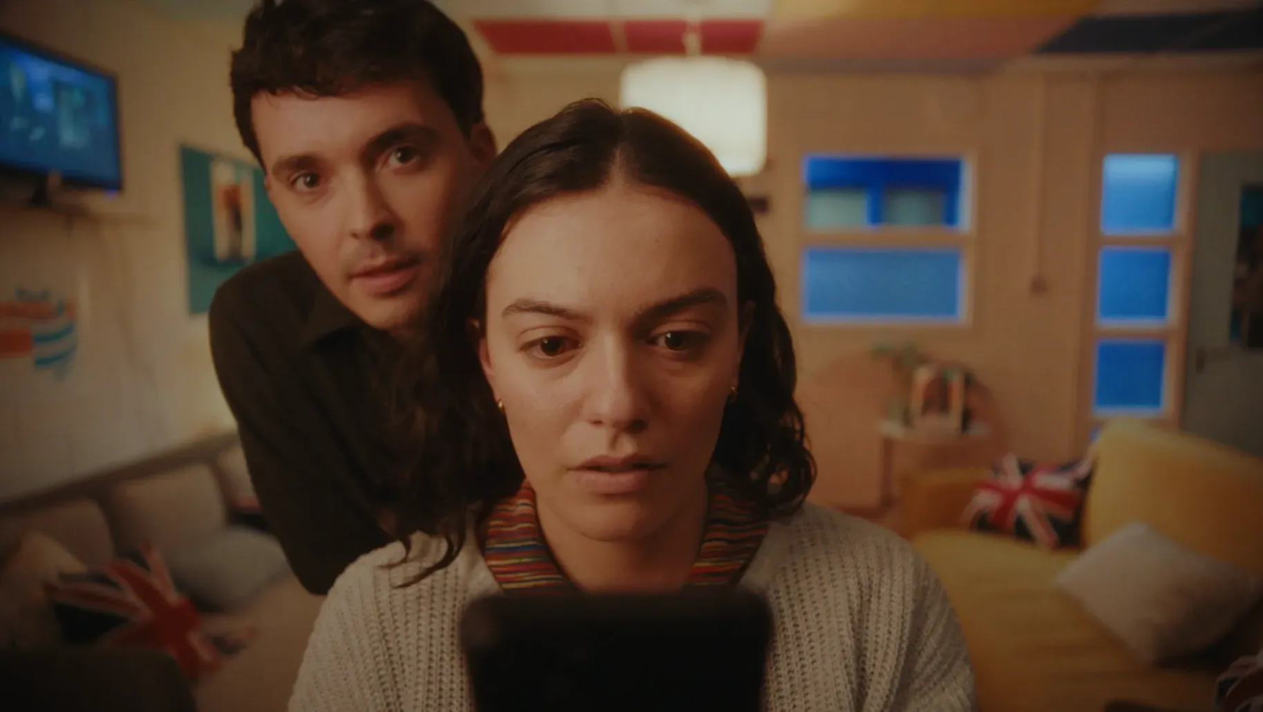 A woman is looking at a phone while a man leans over her shoulder watching her in the mirror