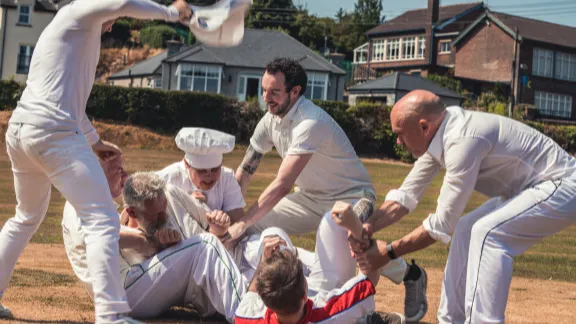 An outdoor skirmish between a group of men in cricket whites and a man wearing a chef's hat.
