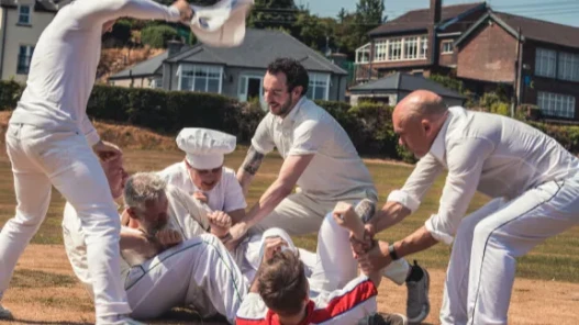An outdoor skirmish between a group of men in cricket whites and a man wearing a chef's hat.