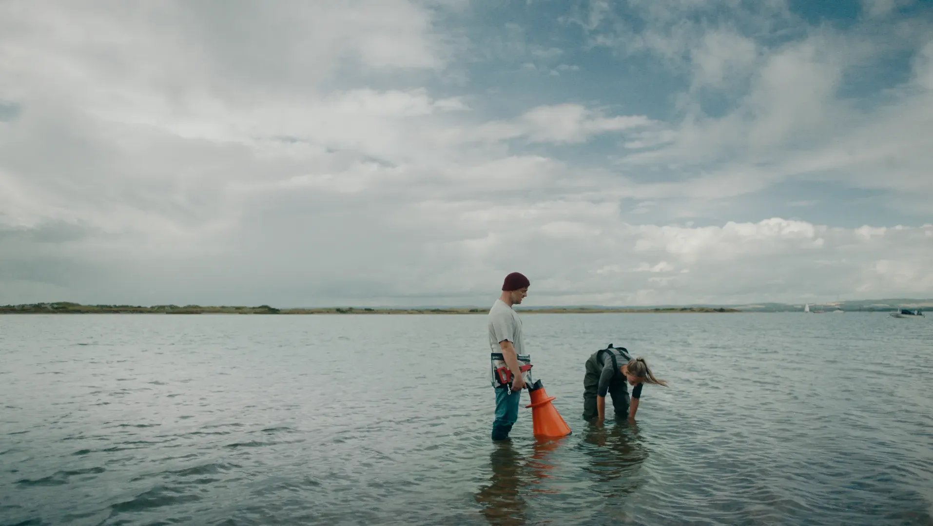 Two figures stand in the sea, one is holding a workbook, the other is leaning over looking into the sea on an overcast, grey day. The sea that surrounds them is calm.