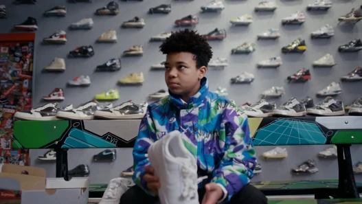 A boy in a colourful patterned jacket sits in a shoe shop holding a white trainer, with rows of trainers displayed on the wall behind him.