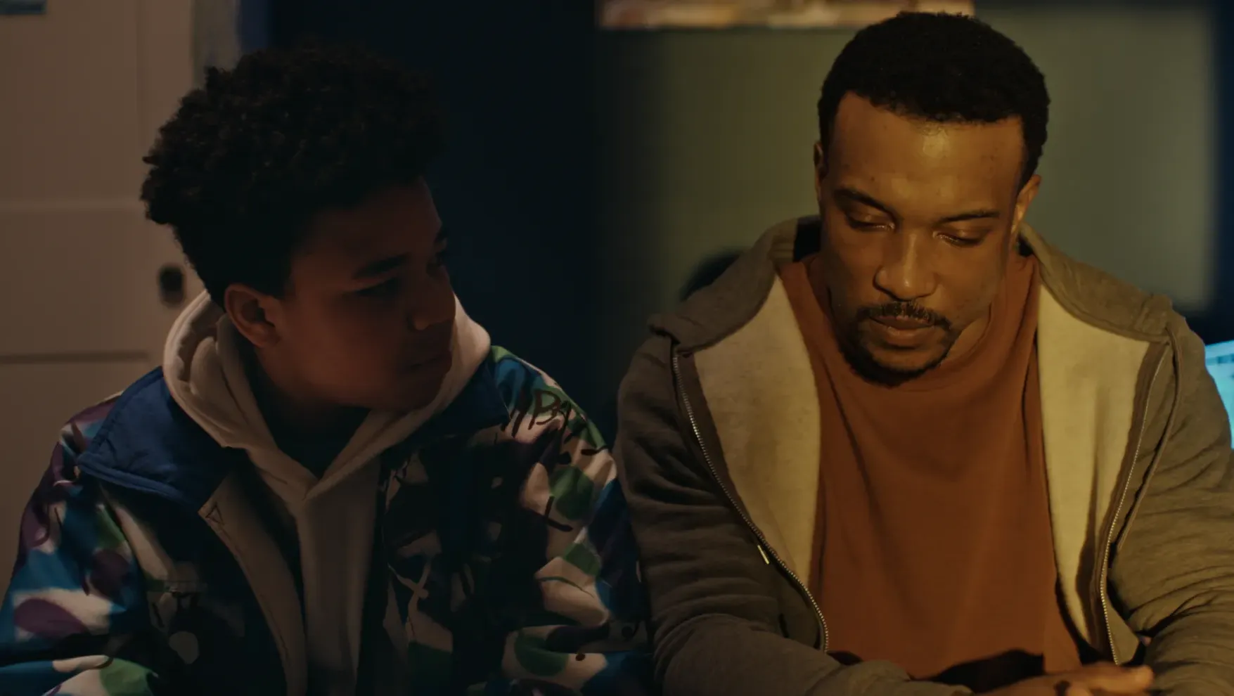 A boy in a colourful patterned jacket sits beside a man in a hoodie and t-shirt, who looks down with a serious expression in a dimly lit room.
