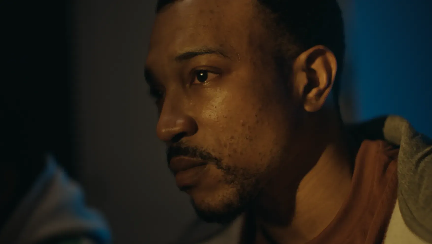 A close-up of a man in a t-shirt and hoodie looking to the side with a serious expression in dim lighting.