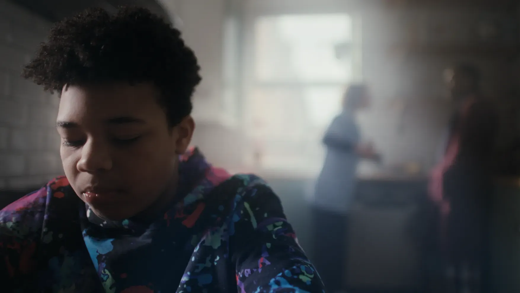 A boy in a colourful patterned sweatshirt looks down with a serious expression in the foreground, while two adults stand blurred in the background of a kitchen.