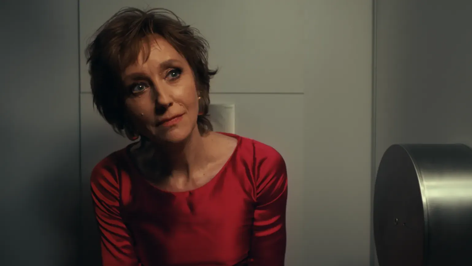 A woman in a red evening gown sits on the toilet in a grey stall, listening.