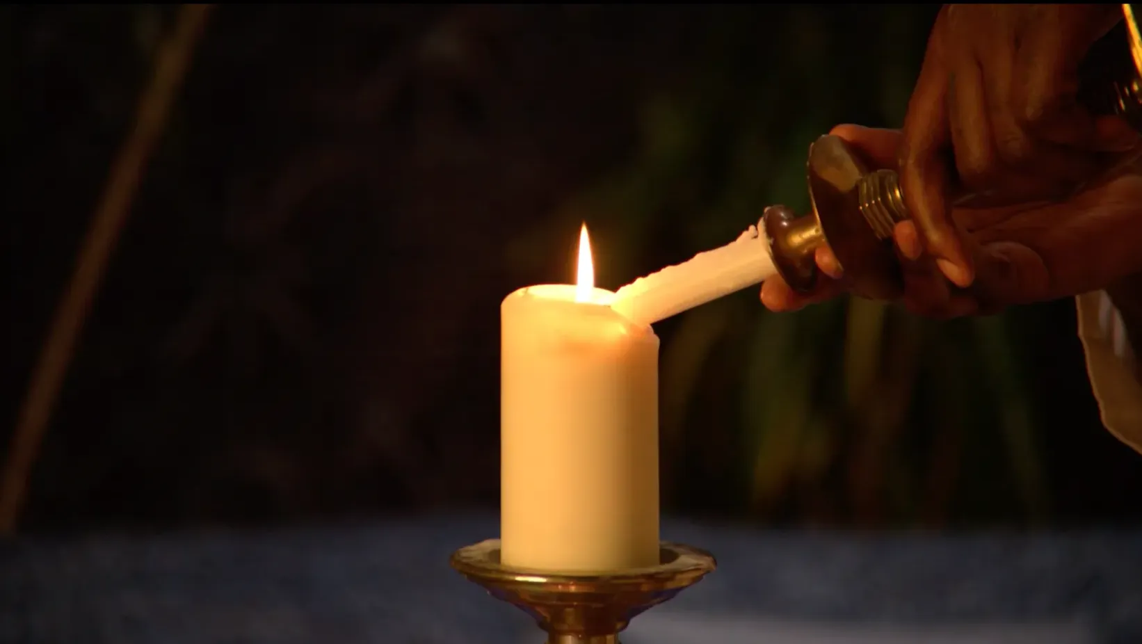 A hand holding a lit taper candle lights a larger pillar candle placed on a metal candle holder, with a dark blurred background.