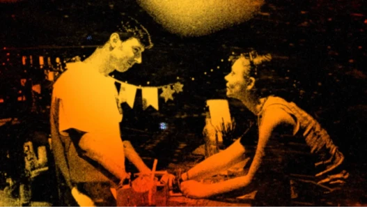 A boy and girl, both 20-something, stare into each other's eyes while touching hands in a dark bar. The image is high contrast, grainy, and tinted orange.