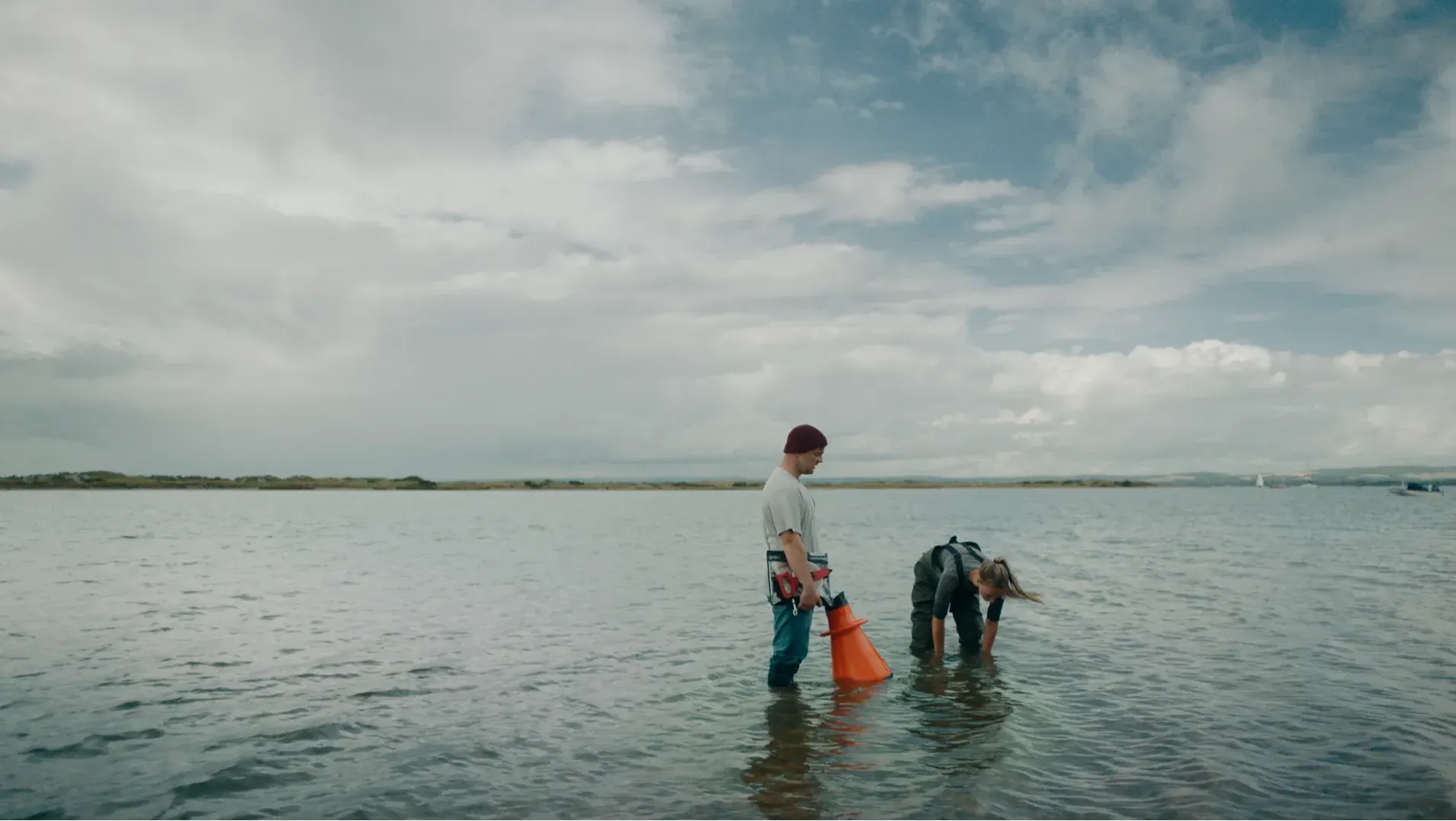 Two figures stand in the sea, one is holding a workbook, the other is leaning over looking into the sea on an overcast, grey day. The sea that surrounds them is calm.