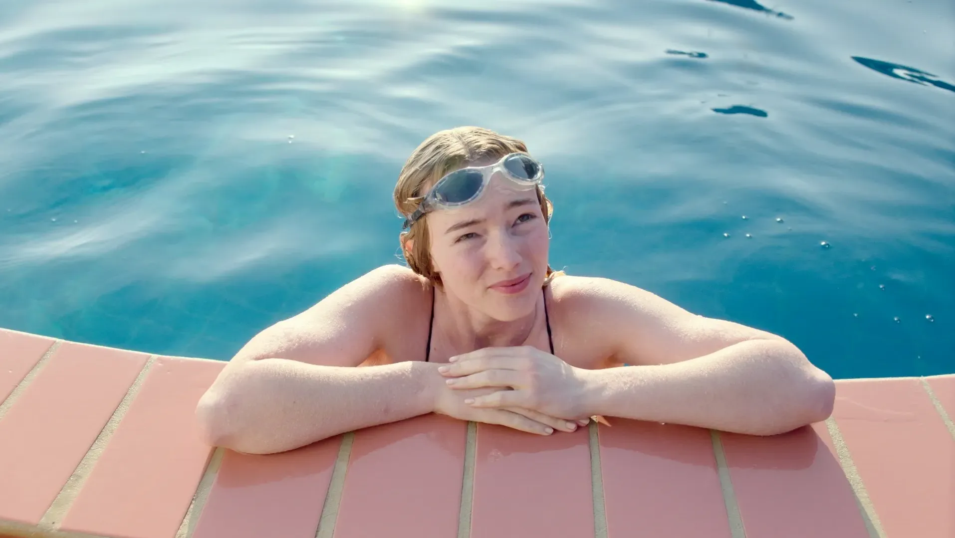 A young woman leans on the side of the pool smiling.