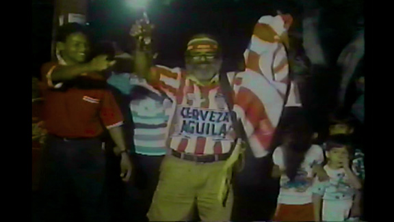 Still from JUNIOR TU PAPÁ (2024) of football fans celebrating in the street.