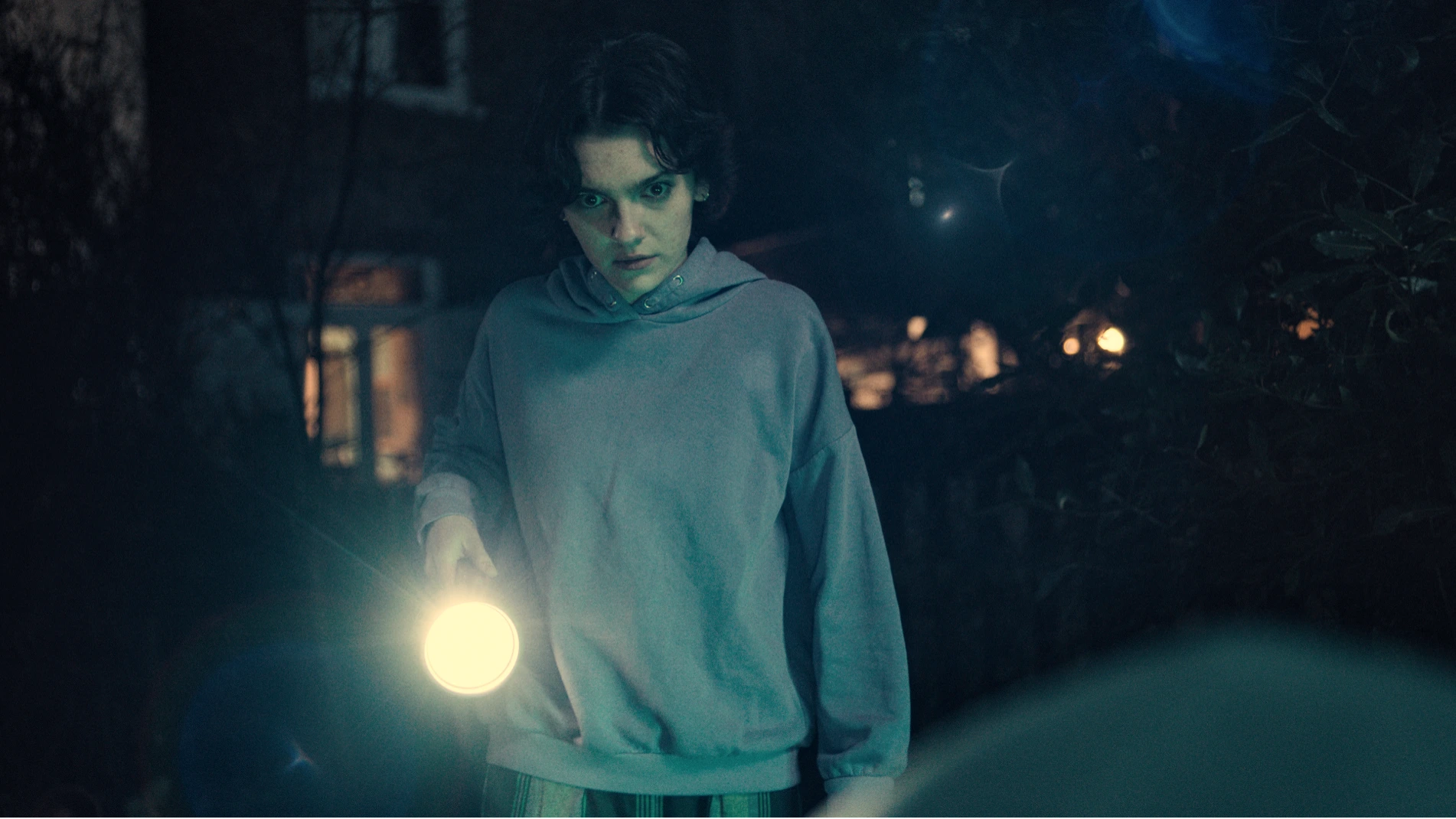 May is in a garden at night, holding a torch at a mysterious egg shaped object in the foreground.