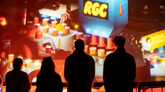 Factory Room at Marshmallow Laser Feast's show Sweet Dreams. People stand by consoles with buttons by a large projection screen depicting a factory.