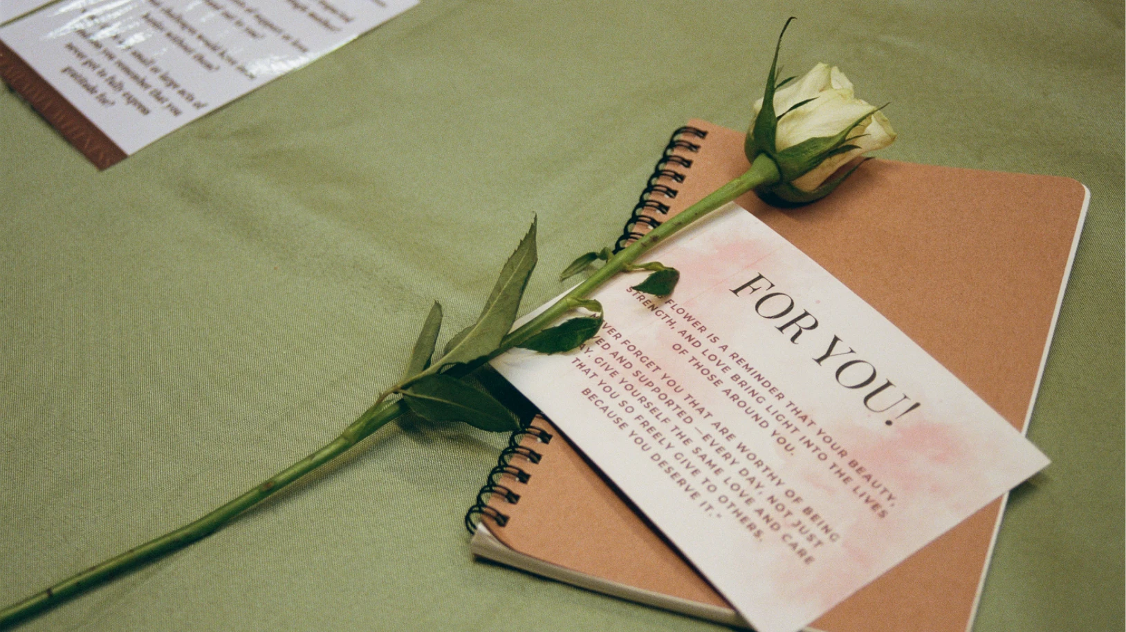 A single white rose sits on top of a card and a brown notebook
