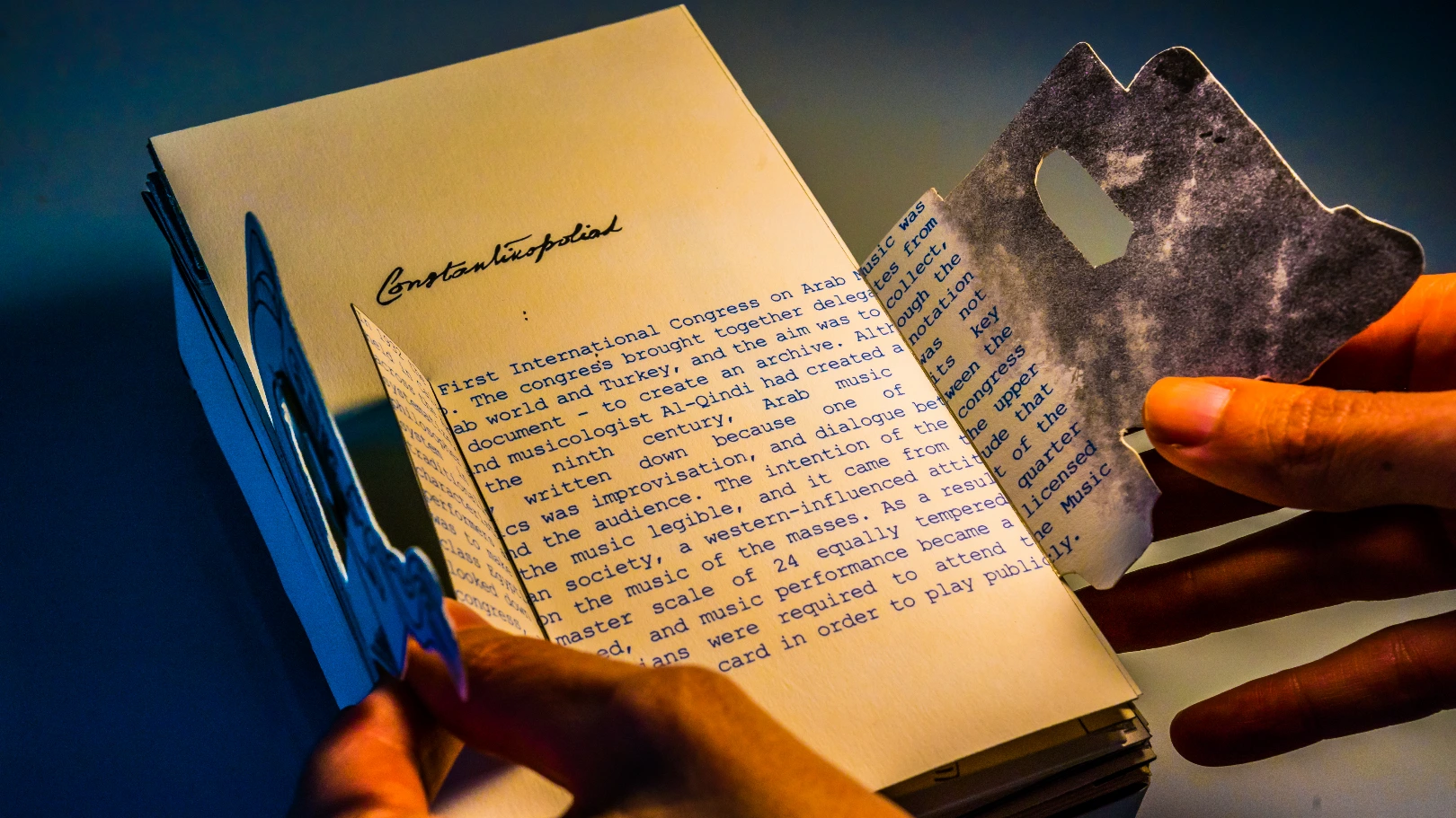 Hands opening the small cover pages of a book, with the title 'Constantinopoliad' above, and text revealed within the pages.