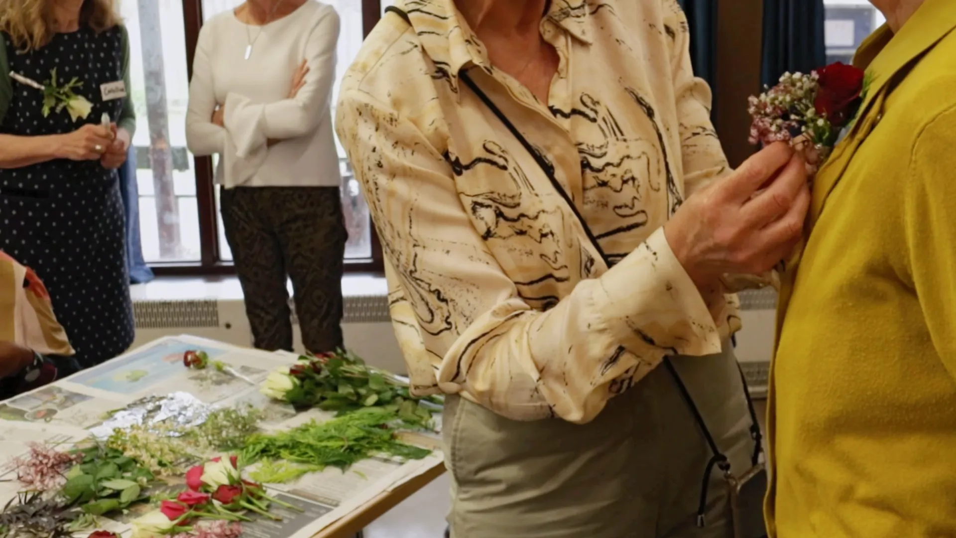 In the foreground, a woman pins a corsage to another woman's cardigan. In the background, two women are chatting in front of a table of cut flowers and foliage.