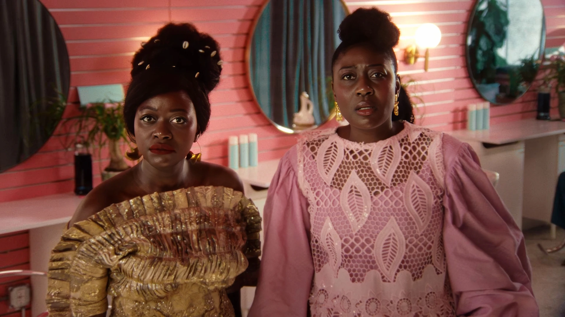 Two Black African women wearing dazzling African native attire, standing in a bright pink beauty salon looking directly at the camera in shock.