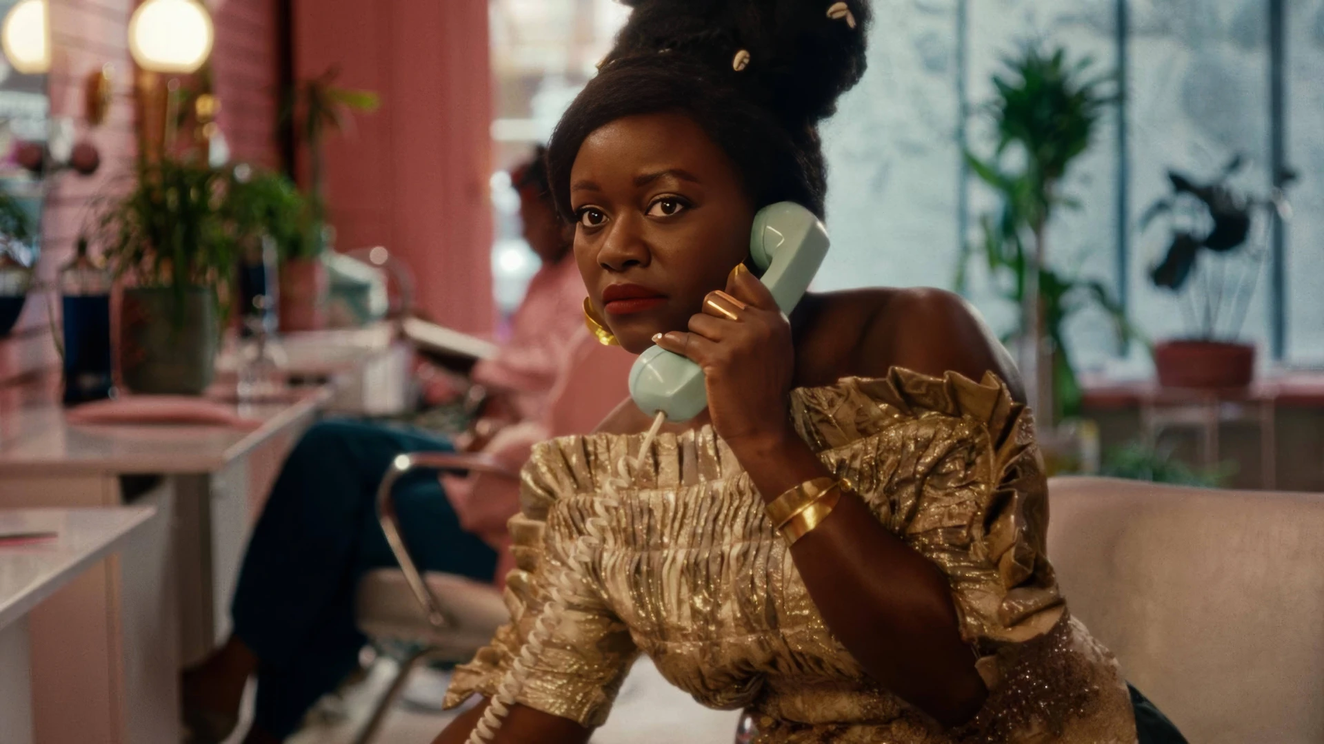 Black African woman wearing a dazzlingly gold top, with a mint green retro hand to her ear, sitting in a hot pink beauty salon.