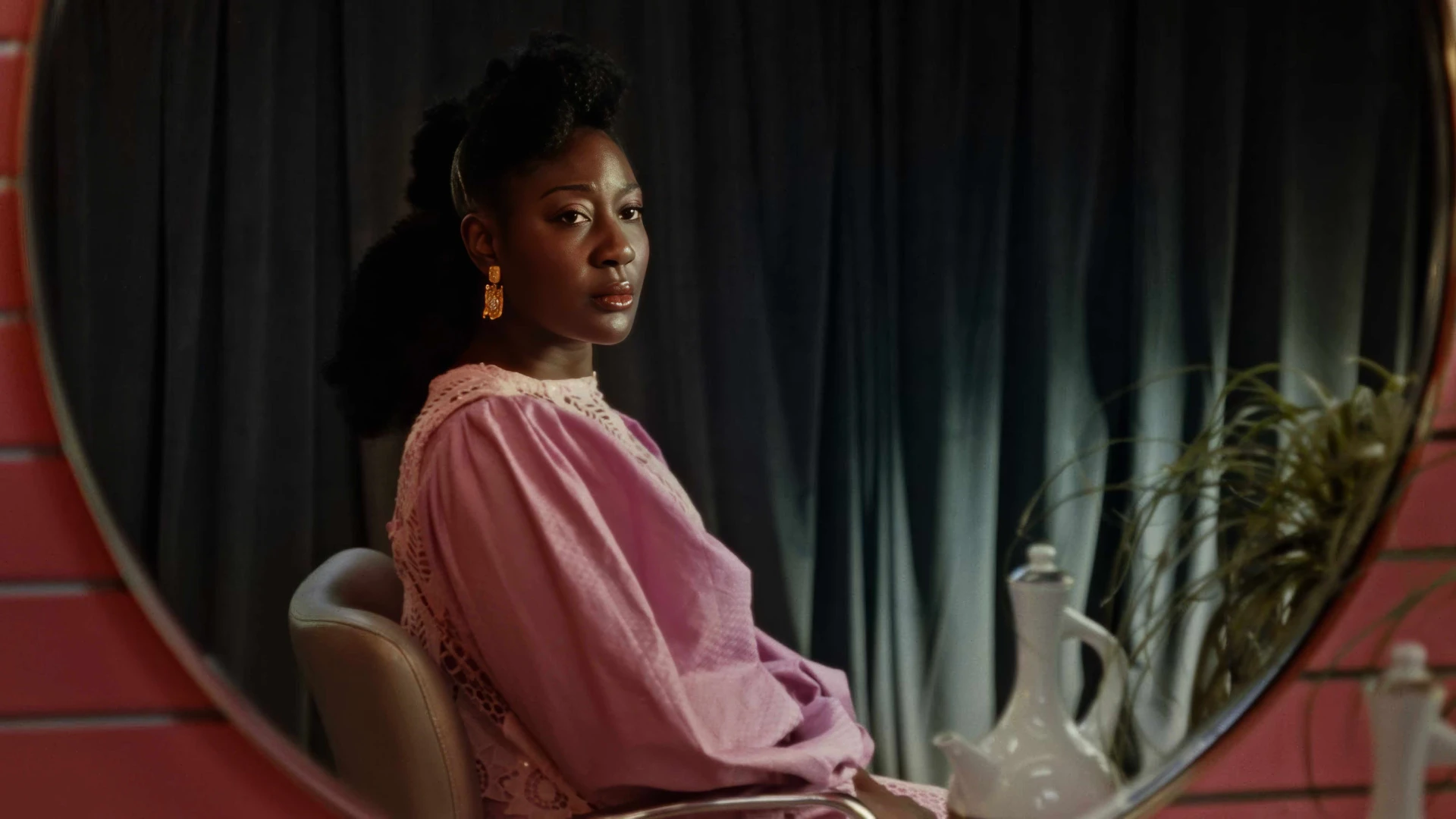 Black African women wearing a dazzlingly pink native outfit sitting, looking at her portrait reflection in a circular mirror.