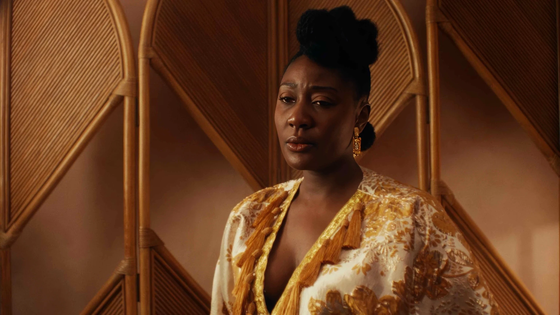 Black African women wearing a dazzlingly gold embellished fringe kaftan in-front of a soft pink wall and a wooden partition. Standing, looking subdued.
