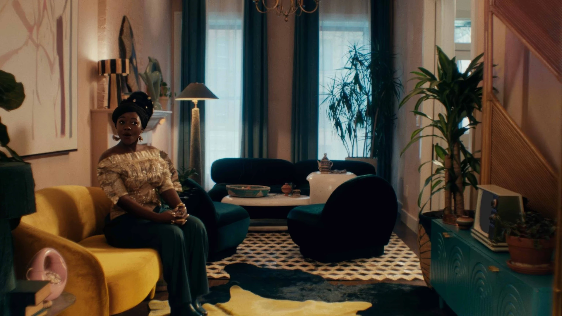 Black African women seated on a gold plush sofa, in a brightly coloured and heavily African inspired patterned living room. With a shocked looked on her face.