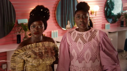 Two Black African women wearing dazzling African native attire, standing in a bright pink beauty salon looking directly at the camera in shock.