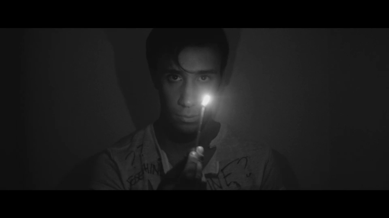 A young man in a dark room holding a lit match and looking menacingly at the camera