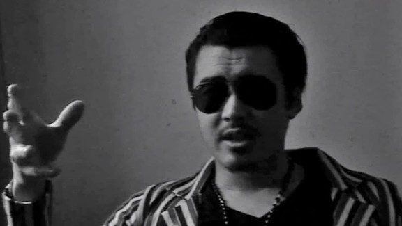 A black and white image of a man wearing sunglasses speaking to someone off camera