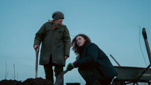 Linda Bassett stands over Charlotte Ritchie, holding a gardening shovel. Charlotte Ritchie is kneeling, also holding a shovel, looking in to the distance. They are planting a hedge.