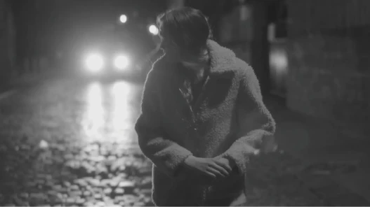 Black and white image. Night time. A woman looks back at a car that might be following her.