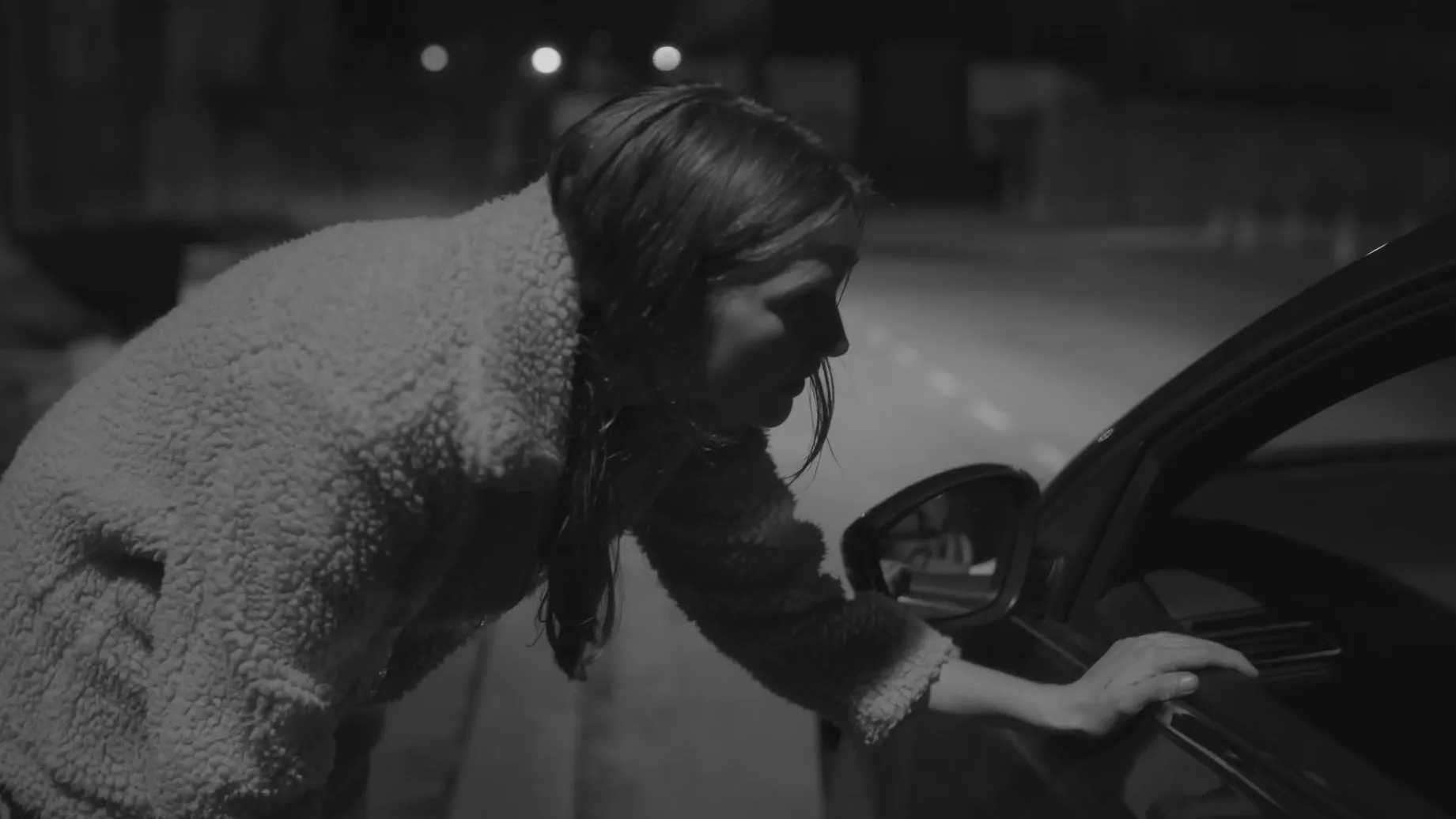 Night time. A woman is leaning down to talk to a driver of a car.