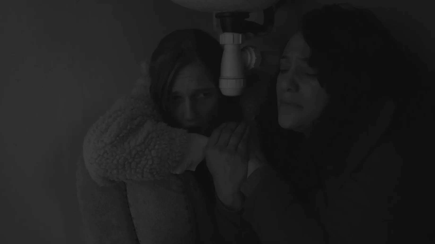 Two women hide under a sink looking afraid.