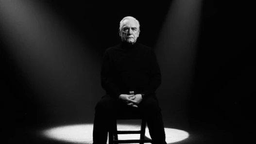 Black and white image. A seated man stares straight into the camera, facing down the audience