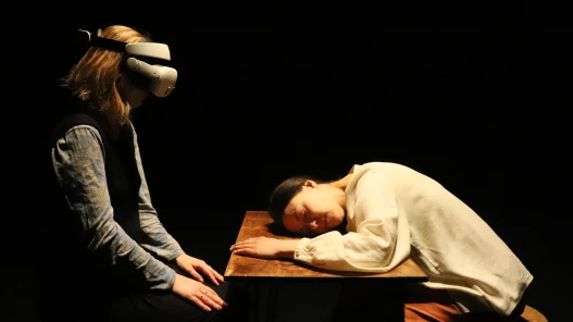 A woman in a dark dress wearing a VR headset is looking at and sitting opposite a woman with dark hair in light clothes with her head down on a dramatically lit table